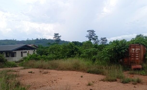 Rural scene with a low building on the left, overgrown grass, and a rusty shipping container on a dirt lot under a cloudy sky.