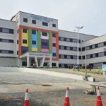 Colorful hospital building with an outpatient entrance, parked car in foreground and orange traffic cones on a paved lot.