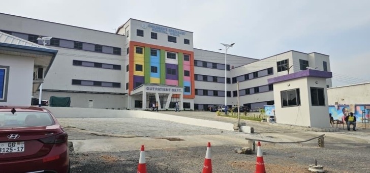 Colorful hospital building with an outpatient entrance, parked car in foreground and orange traffic cones on a paved lot.