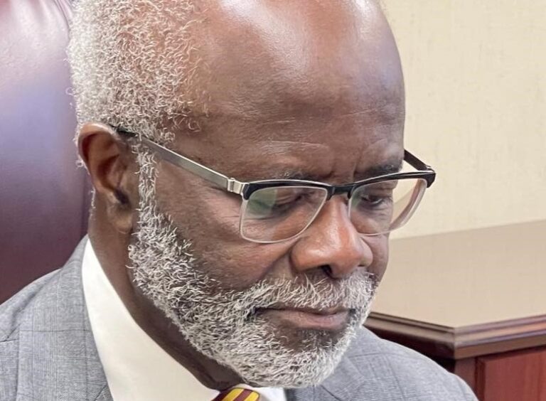 Close-up of an older Black man with a gray beard and glasses, wearing a gray suit, looking down thoughtfully.