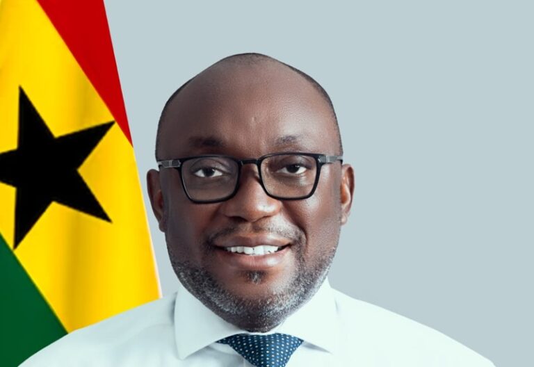 Smiling man with glasses in a white shirt and blue tie, in front of a flag with a black star (Ghana flag).