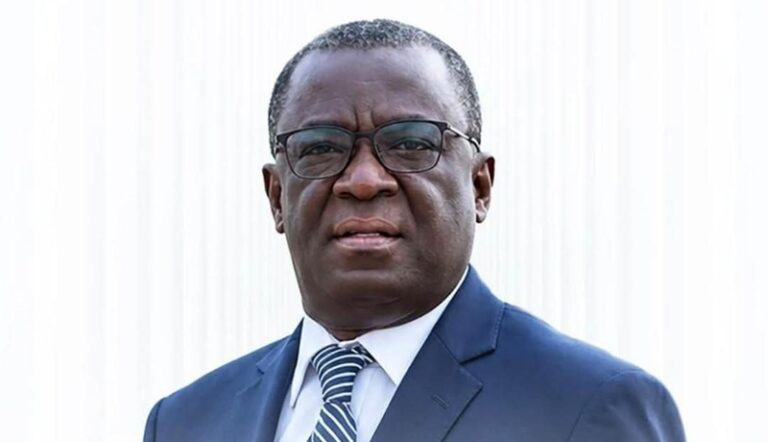 Professional portrait of a middle-aged man wearing a navy suit, white shirt, striped tie, and glasses, looking slightly to the side against a light background.