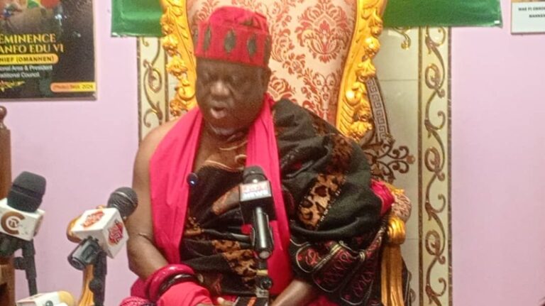 Elderly man in red and black traditional attire sits on an ornate gold chair, speaking at a press conference with multiple microphones in front.