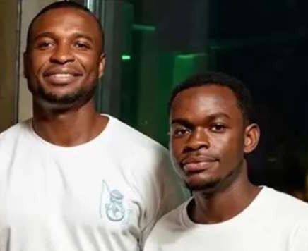 Two men standing side by side indoors, smiling at the camera in white t-shirts with a small logo on the left chest.