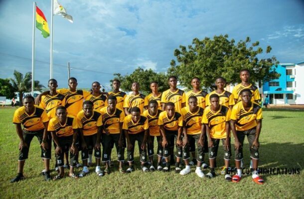 Group of young soccer players in yellow uniforms posing on a field for a team photo with flags in the background.