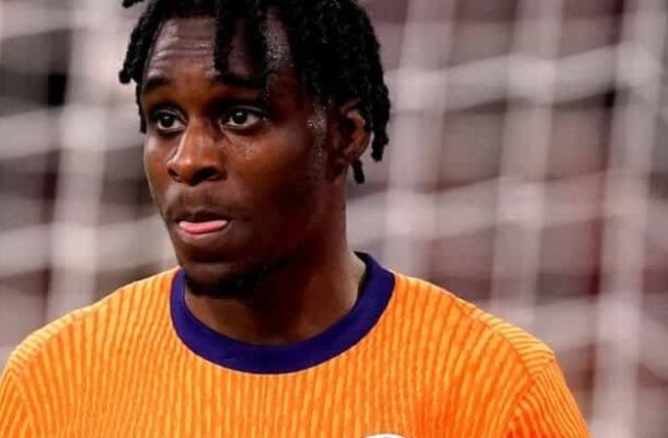 Footballer in bright orange jersey with blue collar, standing on a soccer pitch near a goal.