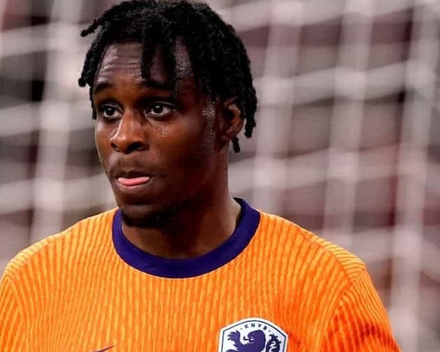 Footballer in bright orange jersey with blue collar, standing on a soccer pitch near a goal.