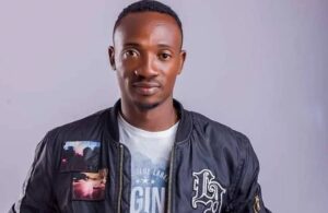 Portrait of a young man facing the camera, wearing a dark bomber jacket with patches over a white graphic tee.