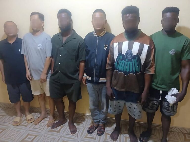 Six men stand in a row against a yellow wall, with faces blurred for privacy.