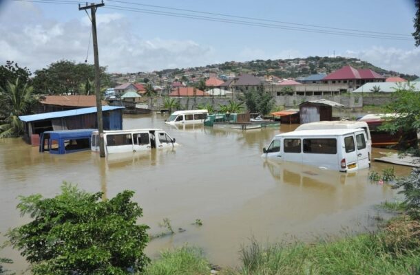 Flooded street in a town with several partially submerged white vans and boat sheds, hillside houses behind.