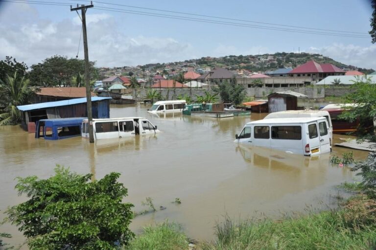Flooded street in a town with several partially submerged white vans and boat sheds, hillside houses behind.