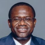 Smiling African man wearing glasses and a navy suit, posing for a formal portrait against a gray background.