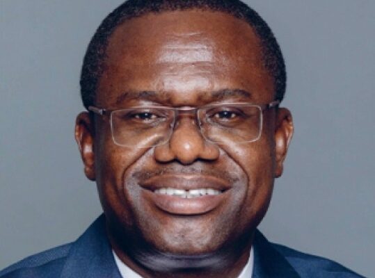 Smiling African man wearing glasses and a navy suit, posing for a formal portrait against a gray background.