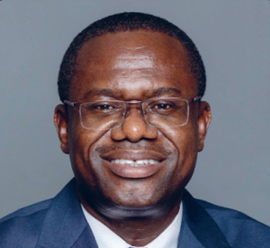 Smiling African man wearing glasses and a navy suit, posing for a formal portrait against a gray background.