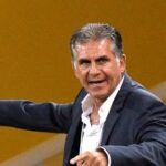 Man in a dark blazer and white shirt extends his arms, pointing as if directing during a event against a yellow background.