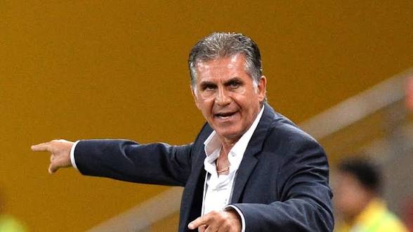 Man in a dark blazer and white shirt extends his arms, pointing as if directing during a event against a yellow background.