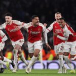 Arsenal players in red and white kits celebrate a goal, running together with arms raised on the pitch.
