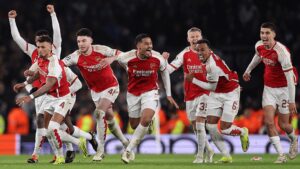 Arsenal players in red and white kits celebrate a goal, running together with arms raised on the pitch.
