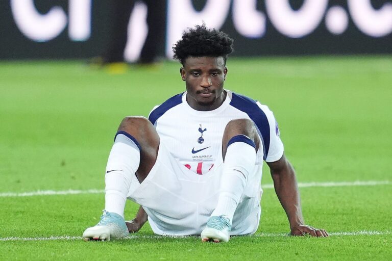 Soccer player in a white and navy kit sitting on the grass with hands on the ground, looking at the camera.
