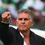 Middle-aged man in a black suit points forward with a serious expression during a sports event on the field.