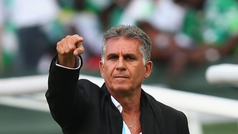 Middle-aged man in a black suit points forward with a serious expression during a sports event on the field.