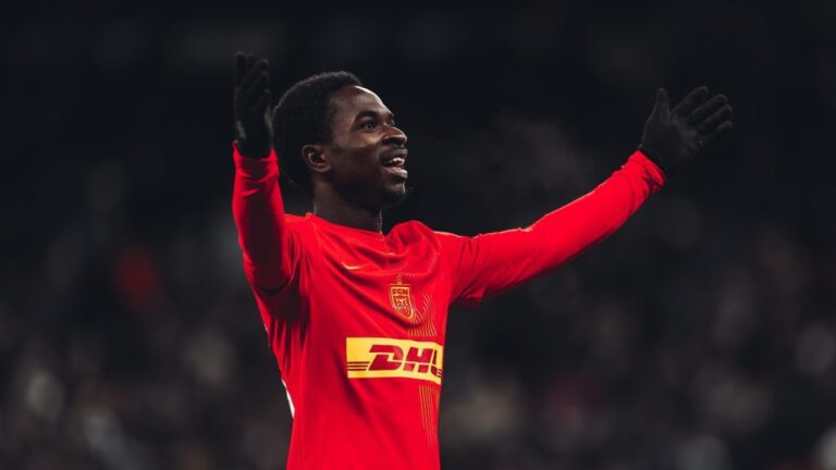 Soccer player in a red jersey raises arms in celebration on the pitch.
