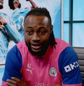 Portrait of a man with a beard and short dreadlocks, wearing a pink Manchester City jersey with blue sleeves and number 42, during an interview or discussion.
