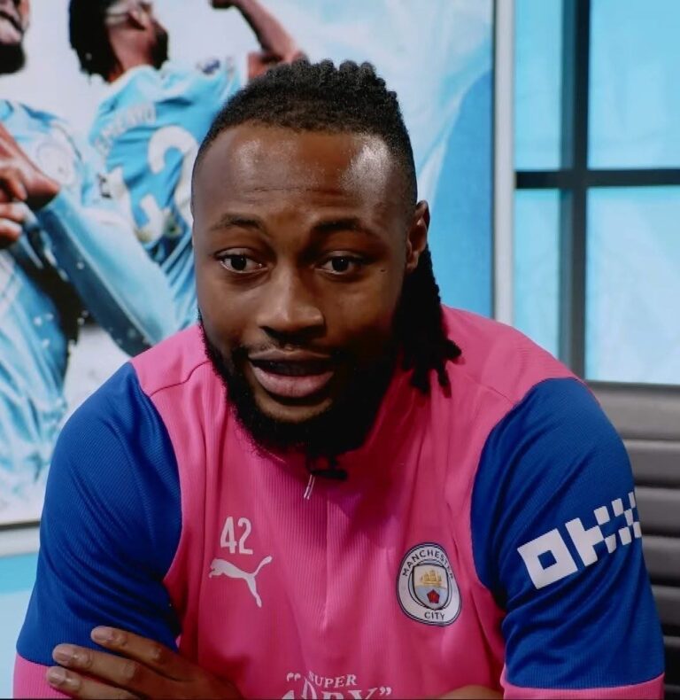 Portrait of a man with a beard and short dreadlocks, wearing a pink Manchester City jersey with blue sleeves and number 42, during an interview or discussion.