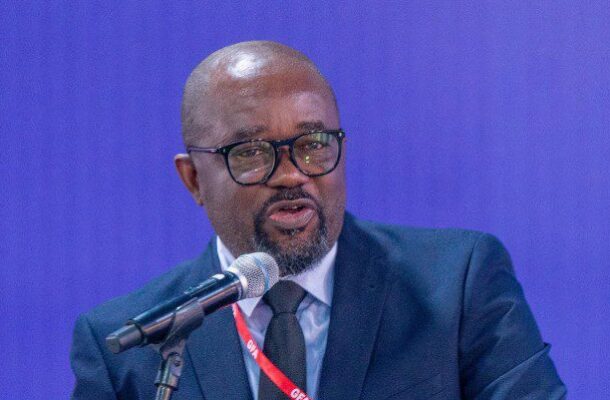 IMG_0259 | Ghanaguardian.com Man in a dark suit and glasses speaks into a microphone at a conference, wearing a red lanyard around his neck.