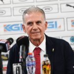 Middle-aged man in a dark suit and red tie at a press conference, speaking to reporters with microphones in front of him and sponsor logos behind.