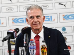 Middle-aged man in a dark suit and red tie at a press conference, speaking to reporters with microphones in front of him and sponsor logos behind.