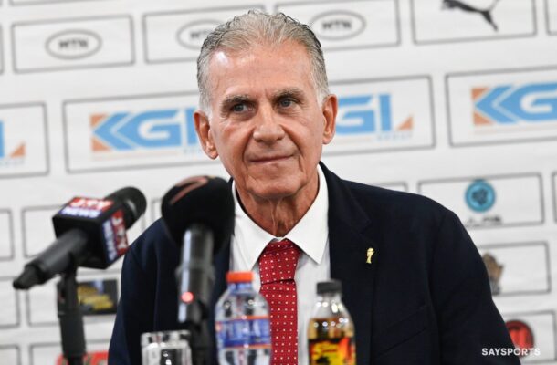 Middle-aged man in a dark suit and red tie at a press conference, speaking to reporters with microphones in front of him and sponsor logos behind.
