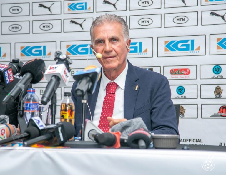Man in a suit and red tie at a press conference, speaking into multiple microphones with a sponsor backdrop behind him.