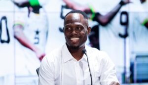 Smiling man in a white shirt speaks into a microphone at a press conference or panel.
