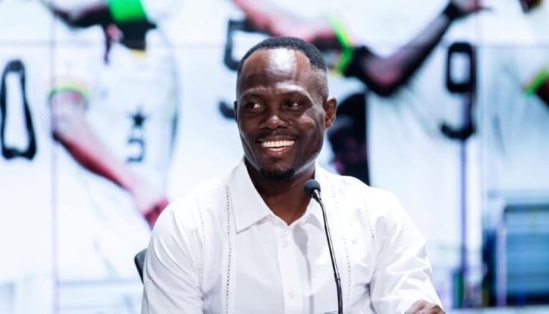 Smiling man in a white shirt speaks into a microphone at a press conference or panel.