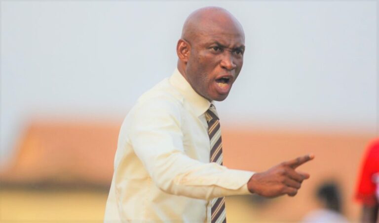 Bald man in a cream shirt and striped tie points and shouts, likely giving instructions or expressing anger on a field.