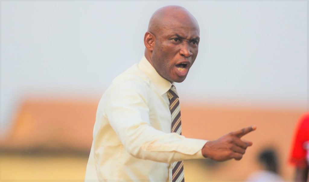Bald man in a cream shirt and striped tie points and shouts, likely giving instructions or expressing anger on a field.