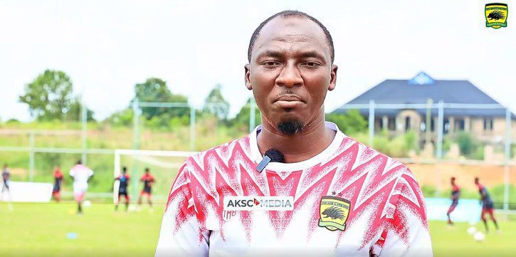 Man in a red-and-white patterned soccer jersey with an AKSC logo speaks to the camera on a sunlit training field, microphone clipped to his shirt.