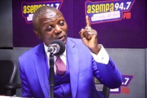 Man in a bright blue suit speaks into a microphone during a radio show at Asempa 94.7 studio, gesturing with one finger raised.