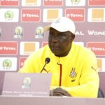 Man in a yellow jacket speaks at a press conference, seated behind a microphone with sponsor logos in the backdrop (CAF/Total/Orange).