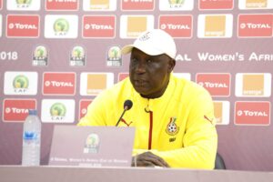 Man in a yellow jacket speaks at a press conference, seated behind a microphone with sponsor logos in the backdrop (CAF/Total/Orange).