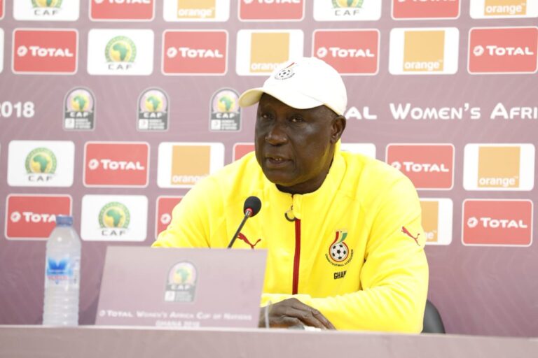 Man in a yellow jacket speaks at a press conference, seated behind a microphone with sponsor logos in the backdrop (CAF/Total/Orange).