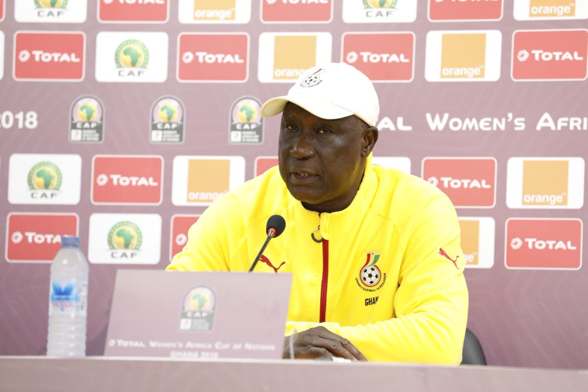 Man in a yellow jacket speaks at a press conference, seated behind a microphone with sponsor logos in the backdrop (CAF/Total/Orange).