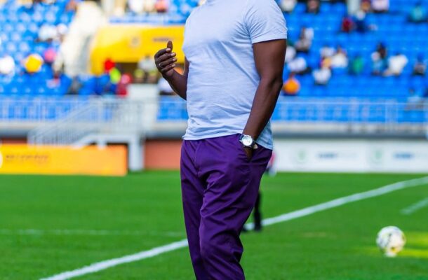 Athlete stands on a soccer field in a white T-shirt and purple pants, with blue stadium seating behind him and a ball at his feet.