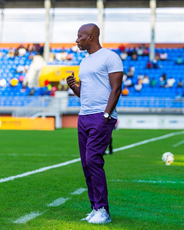 Athlete stands on a soccer field in a white T-shirt and purple pants, with blue stadium seating behind him and a ball at his feet.