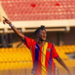 Soccer player in red, yellow, and blue jersey celebrating with arms raised in a stadium stands behind him
