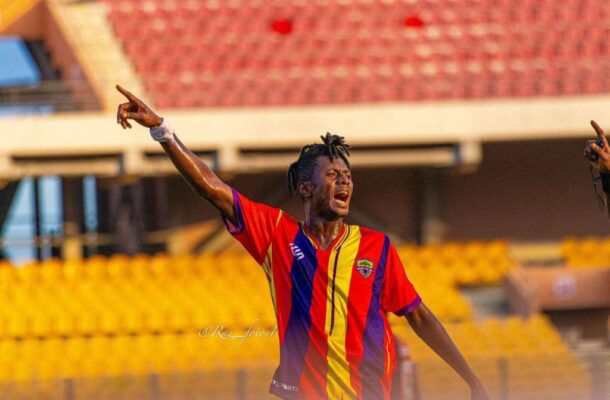 Soccer player in red, yellow, and blue jersey celebrating with arms raised in a stadium stands behind him