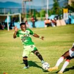 Soccer player in a green jersey (#10) dribbles the ball on a sunny grass field as an opposing player lunges nearby.