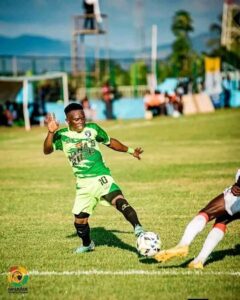 Soccer player in a green jersey (#10) dribbles the ball on a sunny grass field as an opposing player lunges nearby.