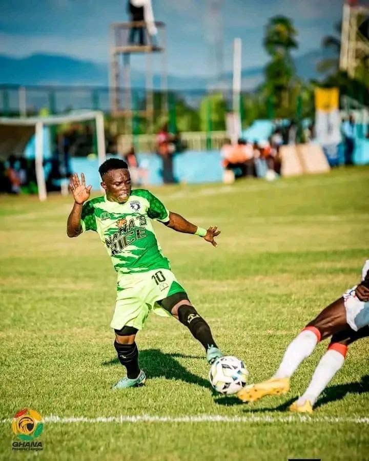 Soccer player in a green jersey (#10) dribbles the ball on a sunny grass field as an opposing player lunges nearby.